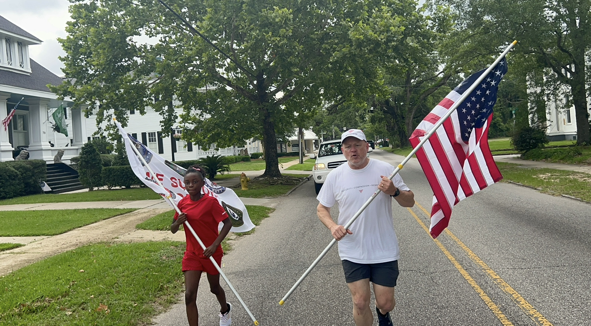 A veteran carrying the American flag runs down a tree-lined Mobile street alongside a young girl carrying a POW/MIA support flag